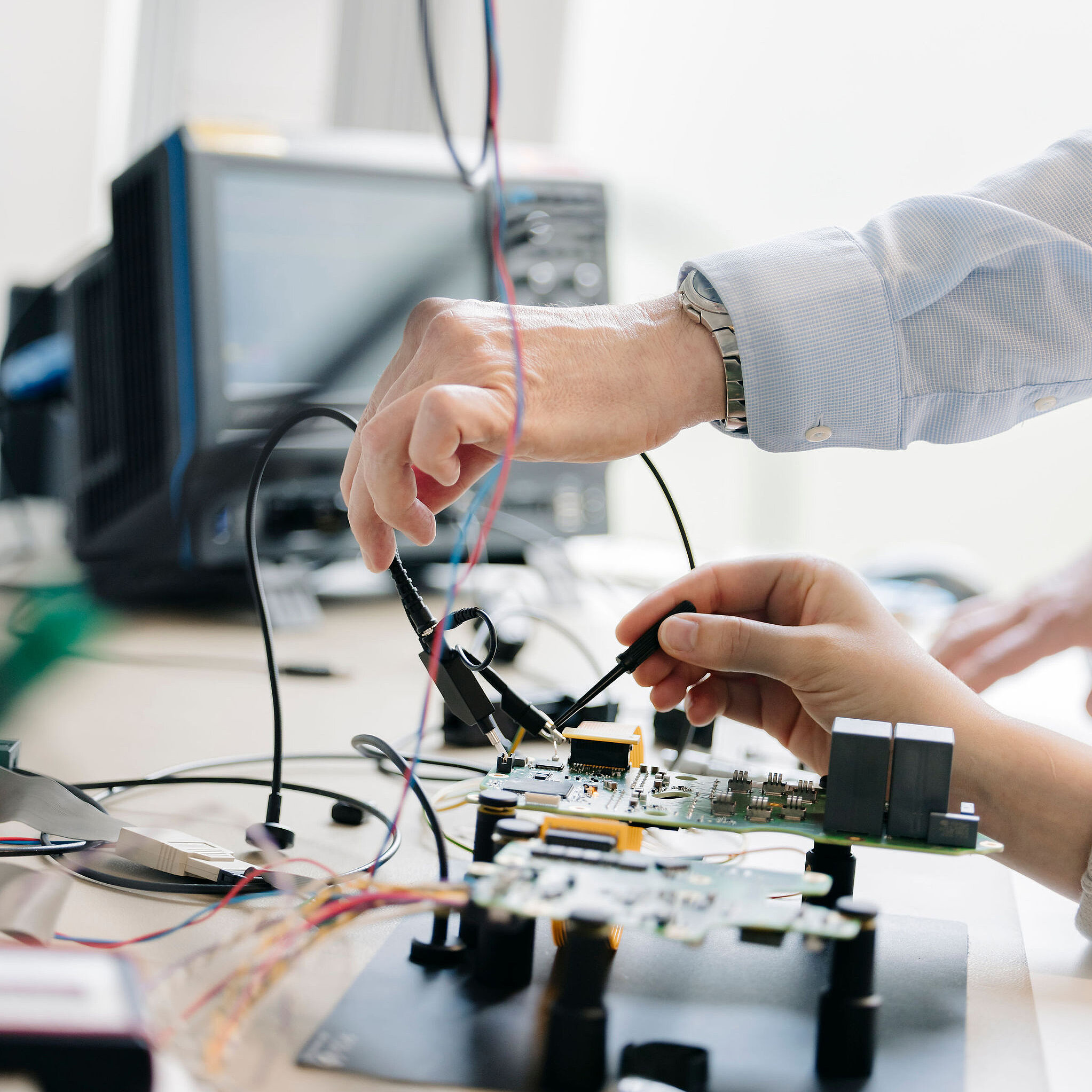 Two Silver Atena employees testing a printed circuit board (PCB) with measurement probes at a laboratory workstation.