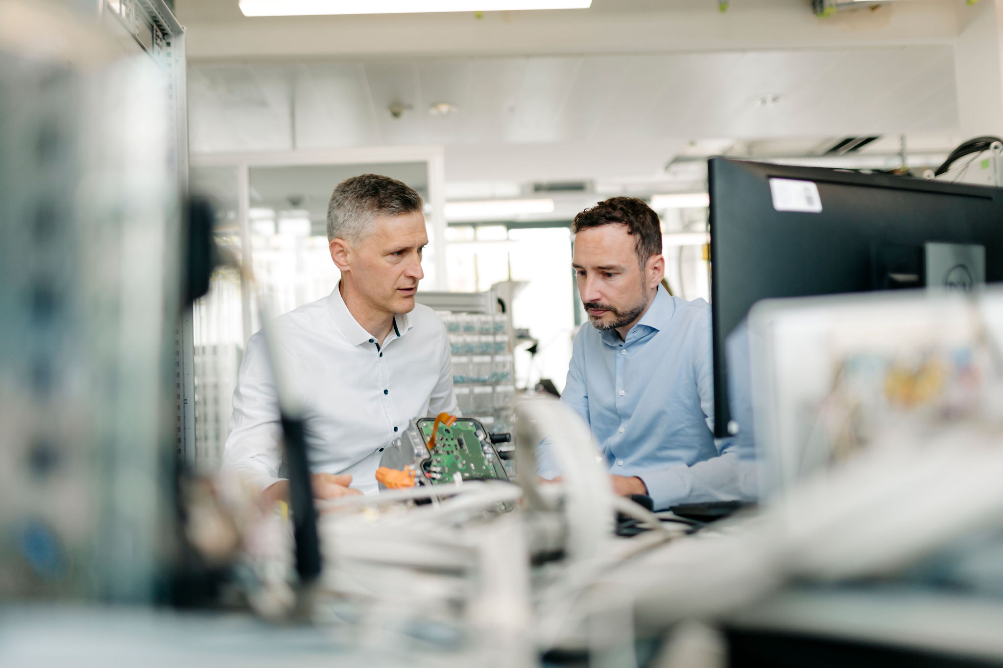 Two engineers examining an electronic assembly with a printed circuit board (PCB) at a development workstation.