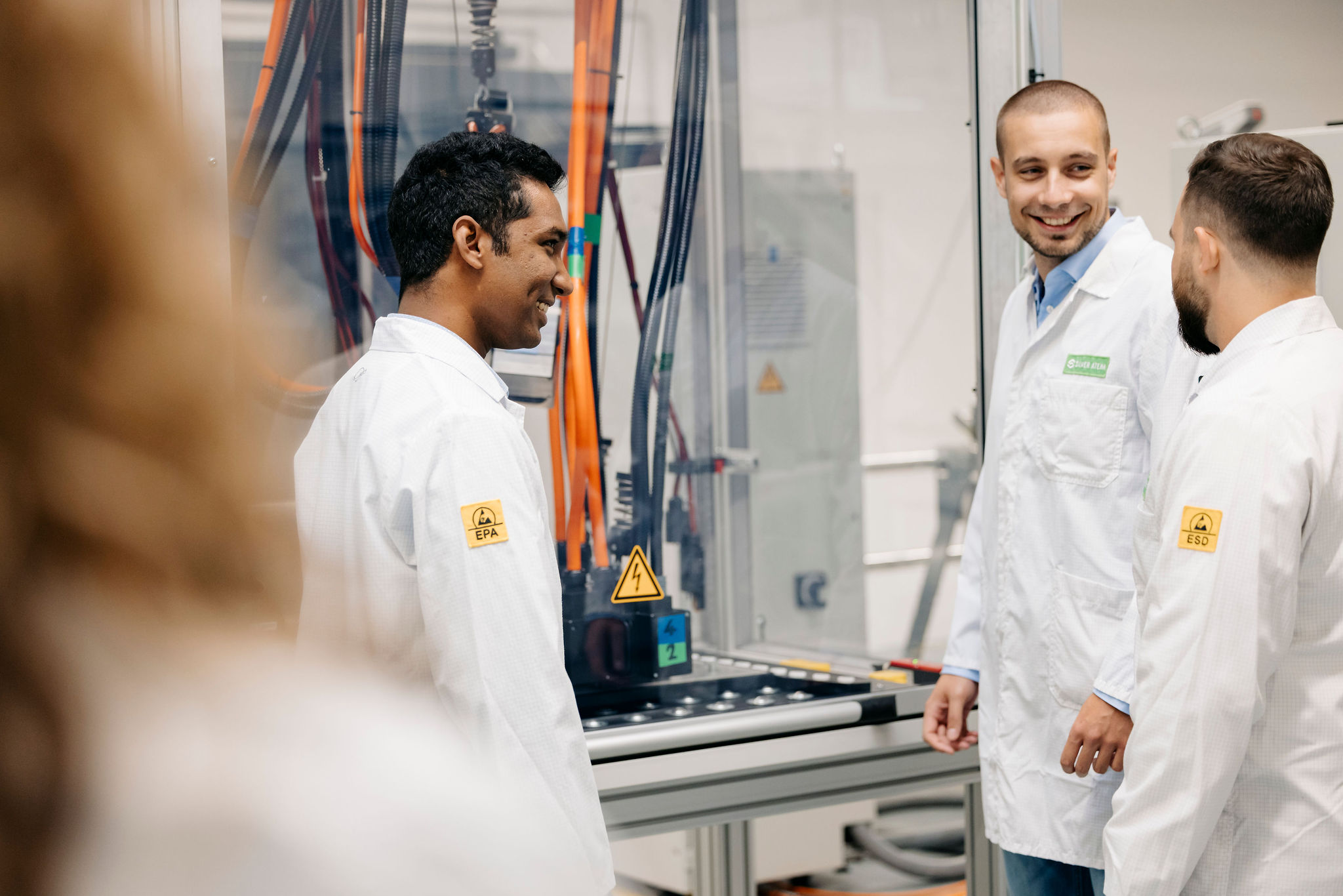 Three Silver Atena employees discussing an industrial test system with high-voltage components and measurement cables.