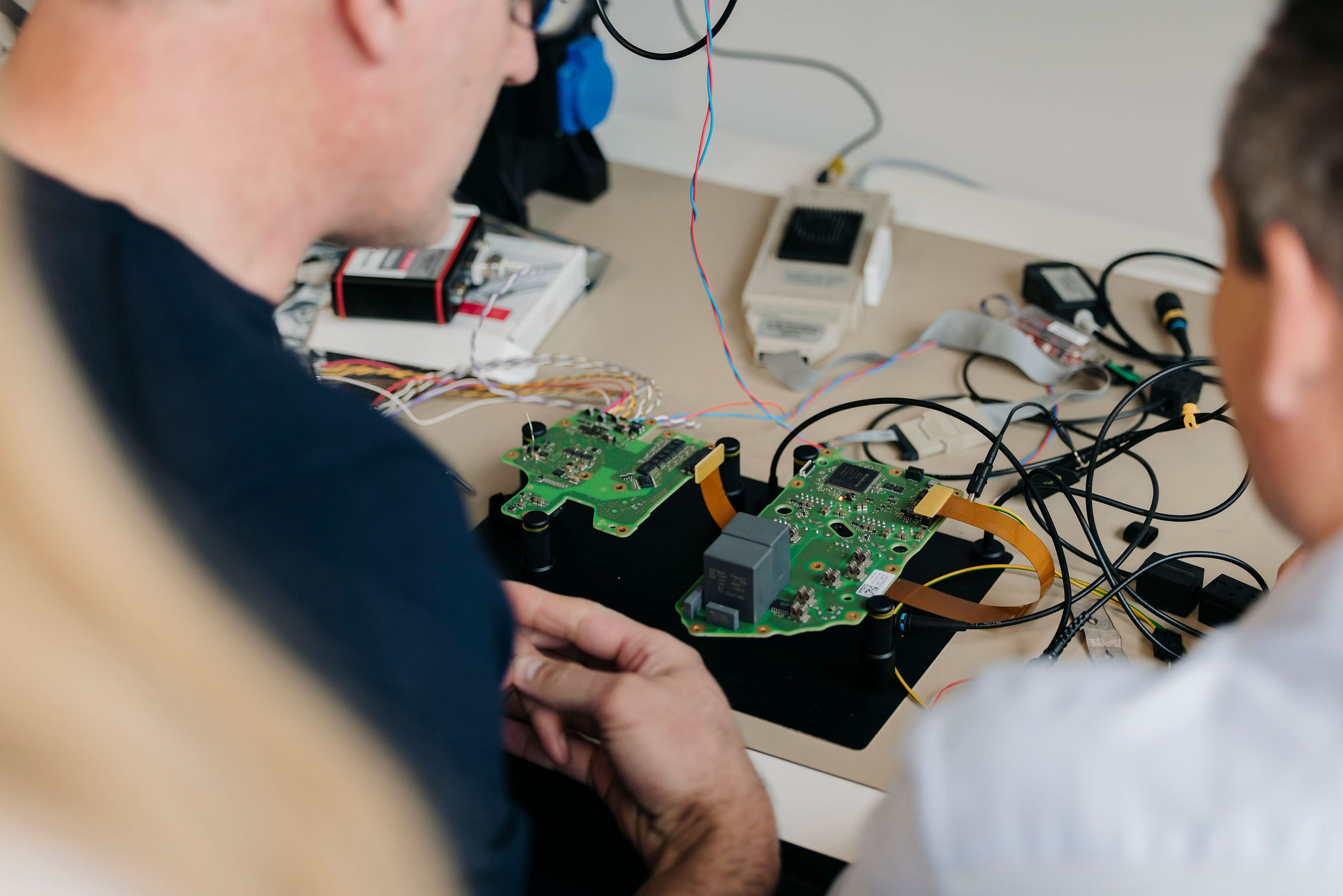 Two Silver Atena engineers examining assembled printed circuit boards (PCBs) with connected measurement cables at a laboratory test bench.